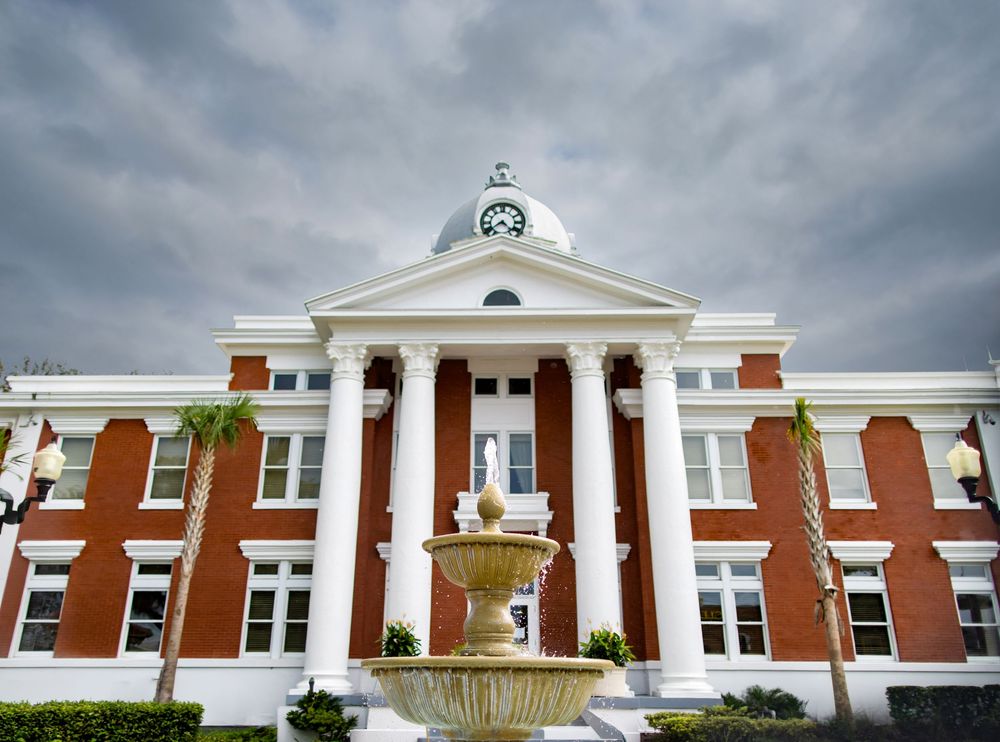 Historic red brick courthouse with white columns, dome clock tower, fountain, and palm trees under dramatic cloudy sky.