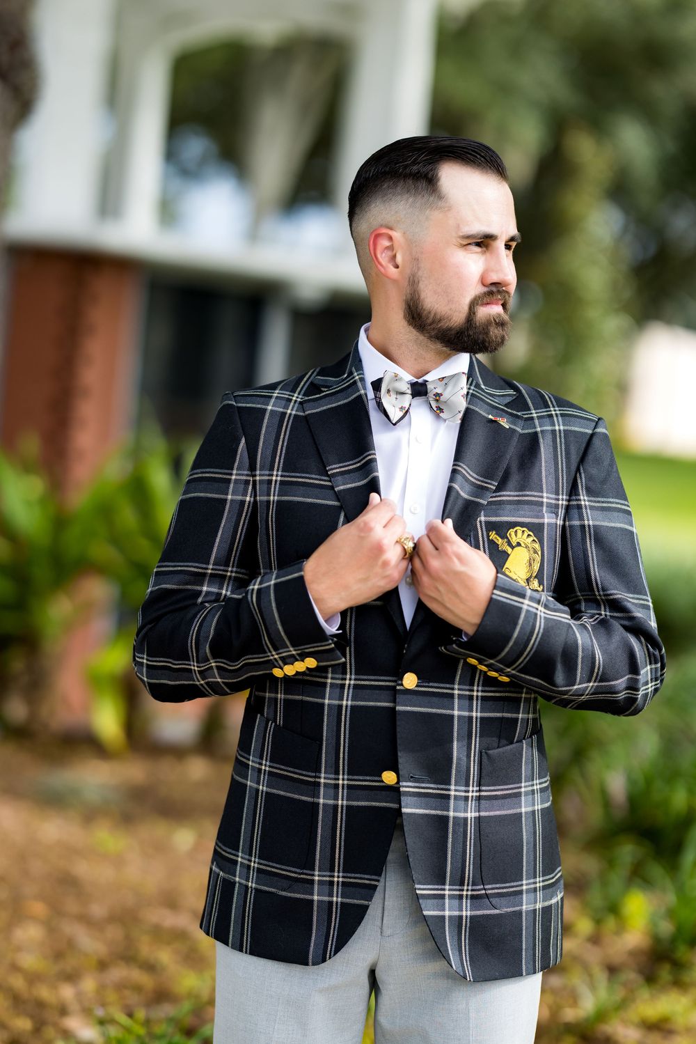 Man in plaid blazer with gold buttons and bow tie adjusting jacket outdoors near building.