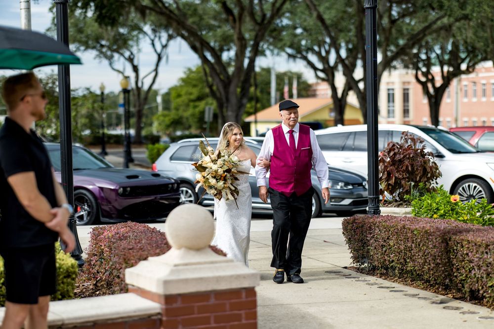 Bride and groom walking together outdoors on a sunny day, bride holding a bouquet, groom in a burgundy vest.