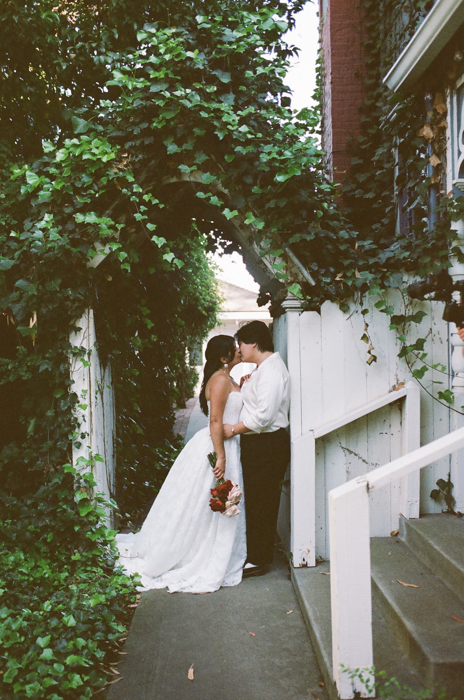 Bride in a white gown holding a red bouquet kisses her groom beneath an ivy-covered arch beside a white picket fence.