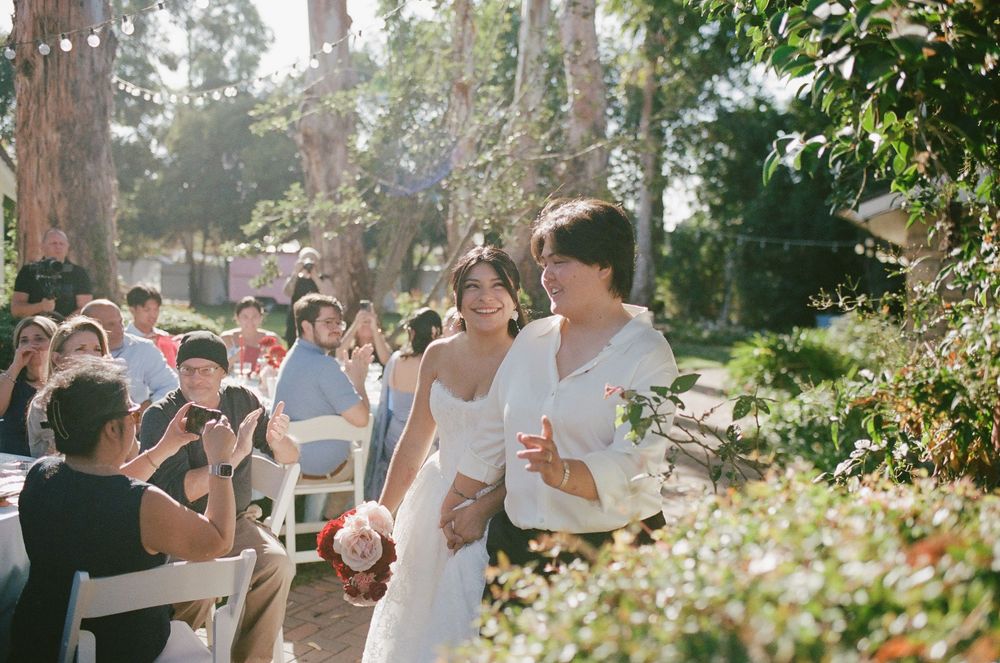 Two brides in white walk arm in arm and laugh down the aisle at an outdoor garden wedding as seated guests cheer beneath tall trees.