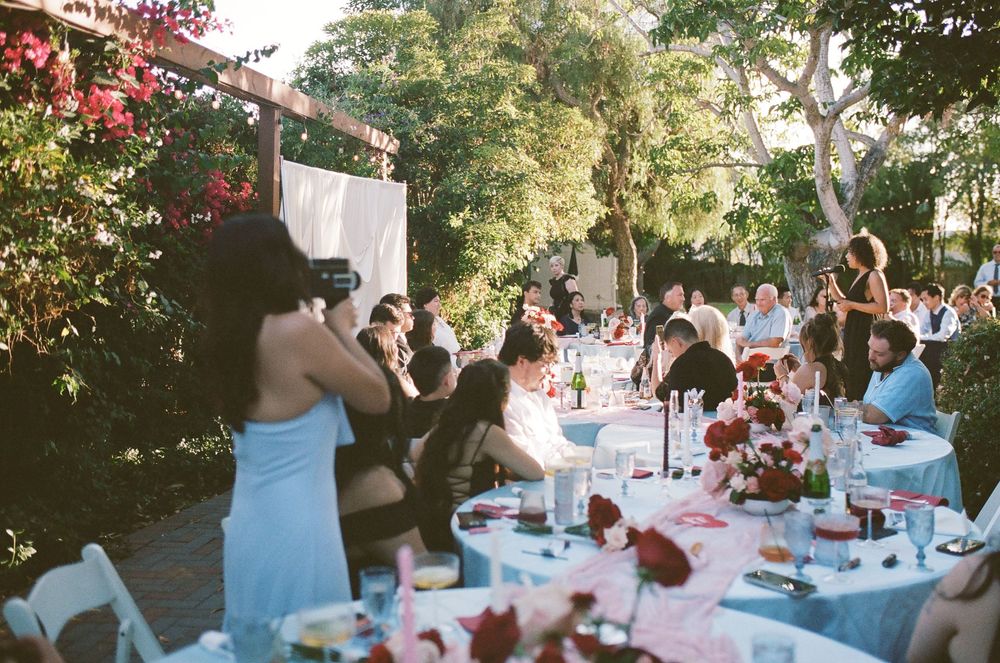 Wedding guests seated at round tables with pink floral centerpieces under shady trees at an outdoor garden reception.