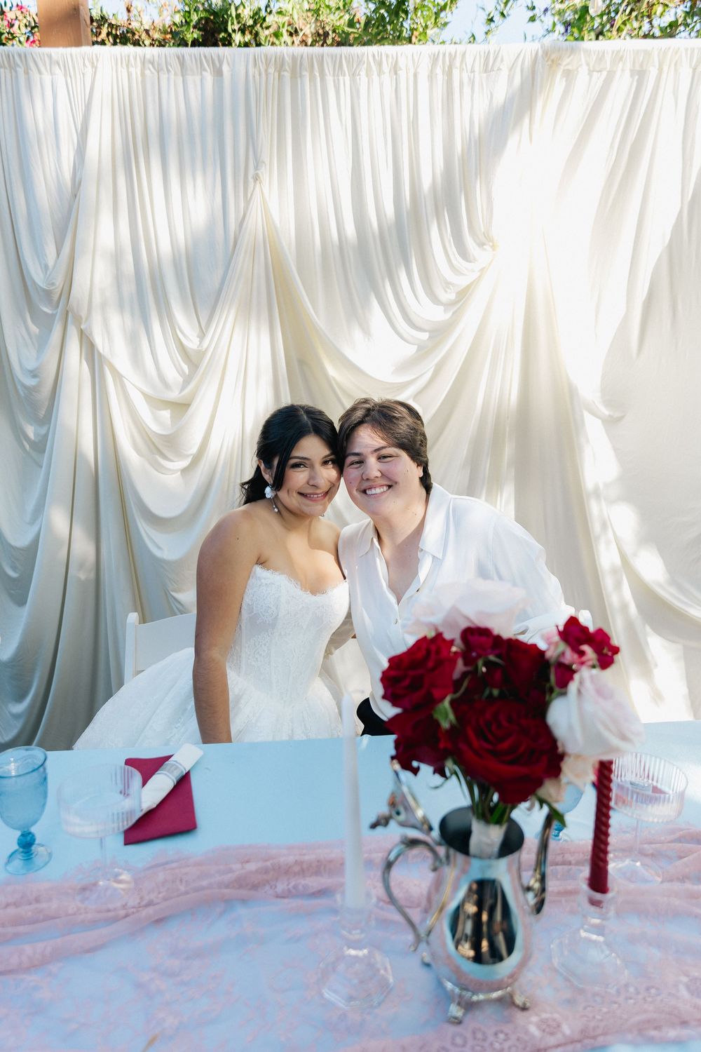 Two smiling brides sit together at a wedding reception table with red roses, a silver pitcher, and white draped curtains behind them.