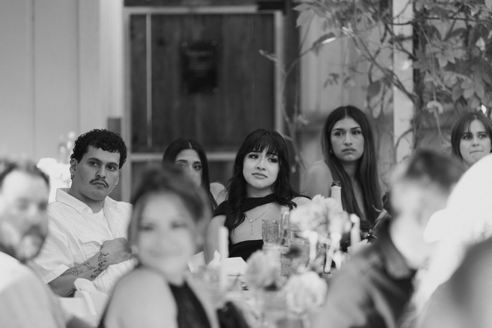 Black and white photo of wedding guests seated at a reception table, watching attentively with flowers and glassware in the foreground.