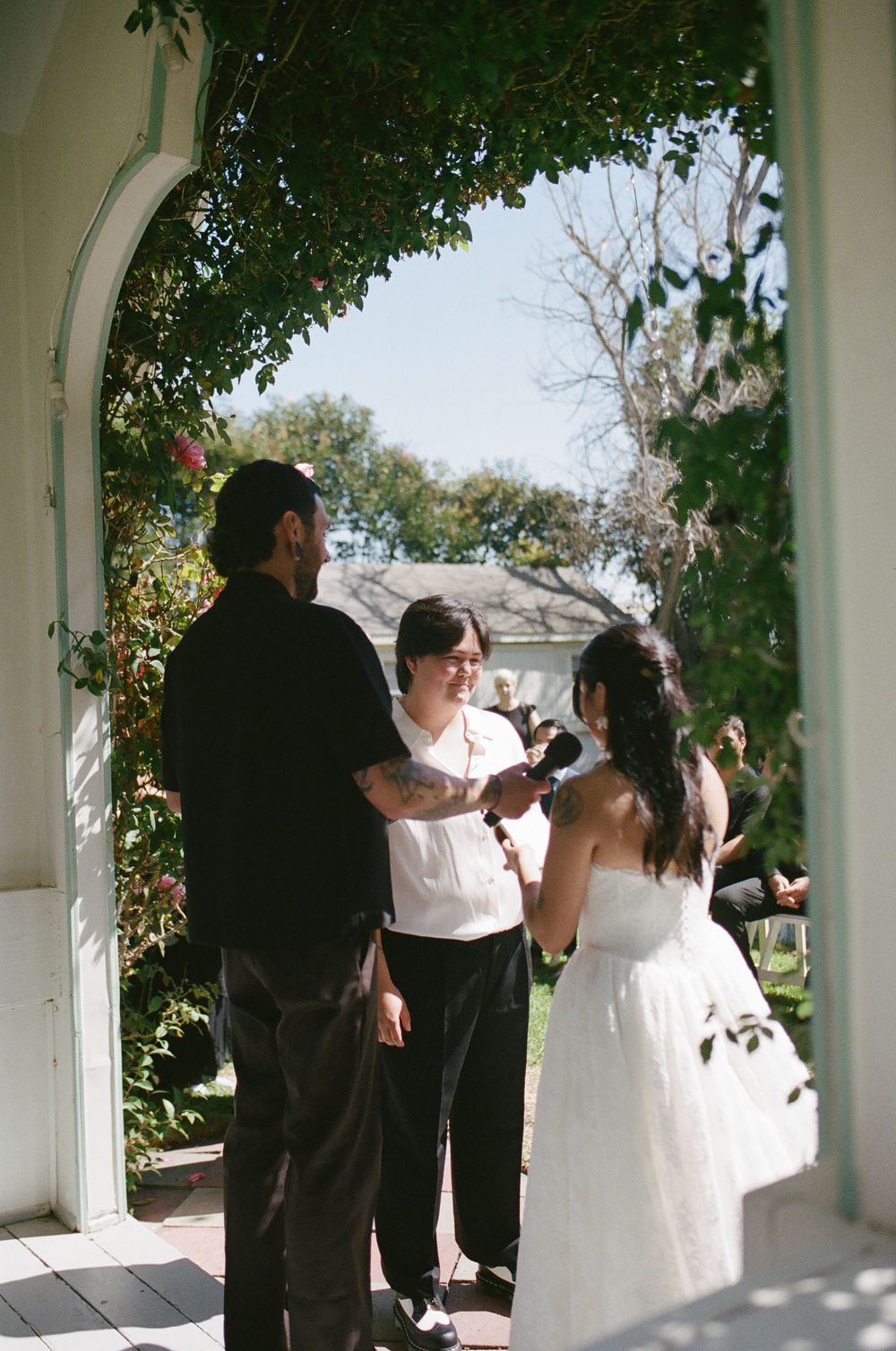 Bride in a white dress and groom exchange vows with an officiant under a flowering garden archway during an outdoor wedding ceremony.
