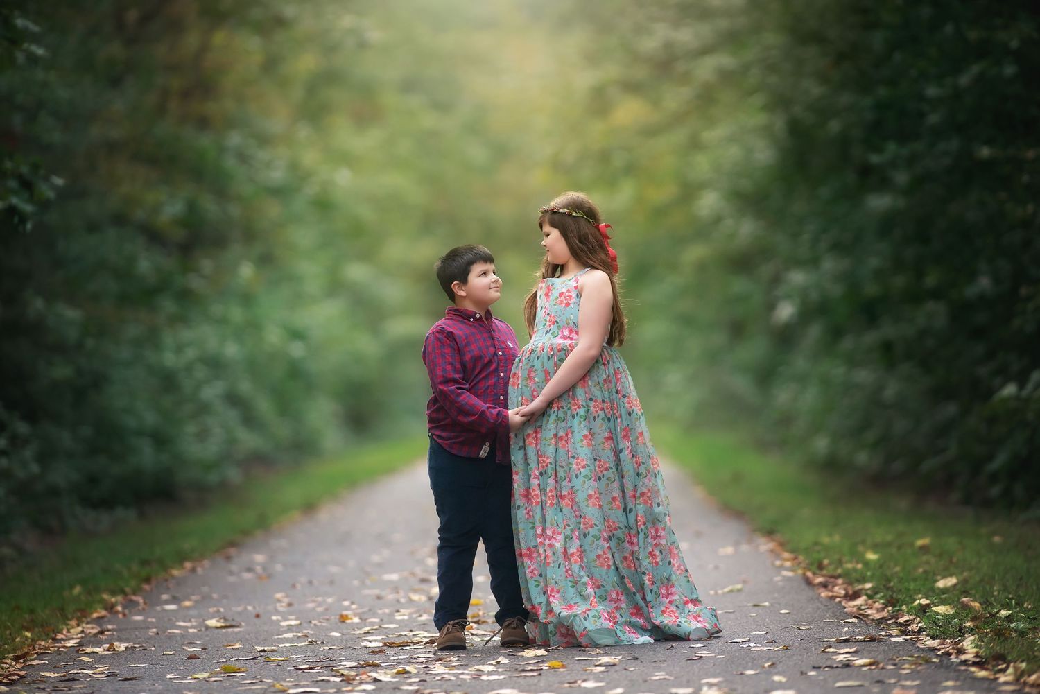 Pregnant woman in a floral maxi dress holds hands with a young boy on a leaf-covered tree-lined path in a maternity photo session.