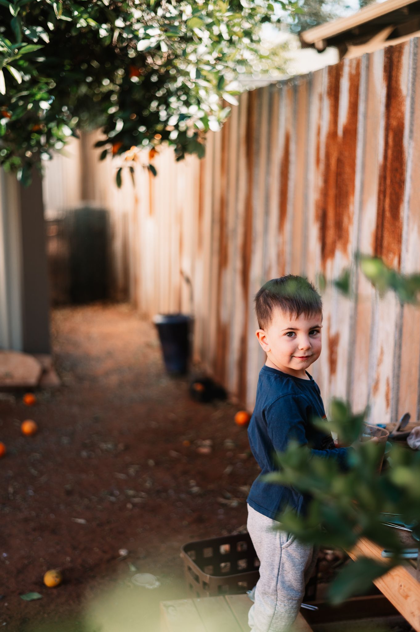Young child in a navy sweater plays at a wooden outdoor mud kitchen in a sunlit backyard with fallen oranges scattered on the dirt.