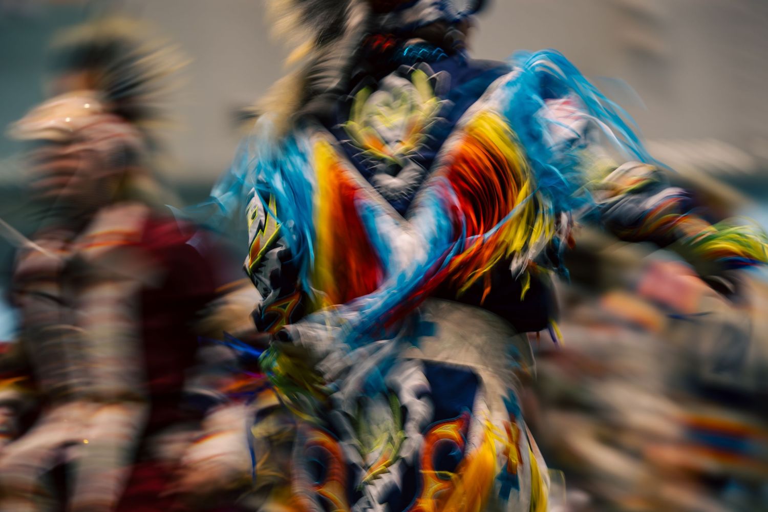 Motion-blurred dancer in vibrant blue, orange, and yellow fringed regalia spinning at a powwow, with a second dancer blurred behind.