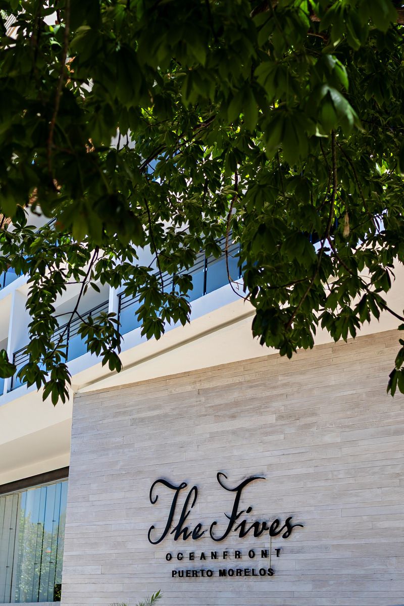 The Fives Oceanfront Puerto Morelos hotel exterior sign on a stone wall framed by lush green tree branches.