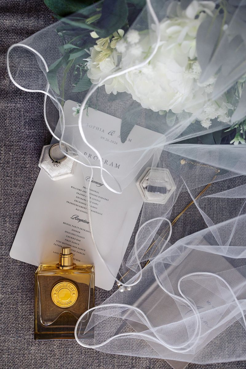 Elegant wedding flat lay with bridal veil, white floral bouquet, program, ring boxes, and gold perfume bottle.