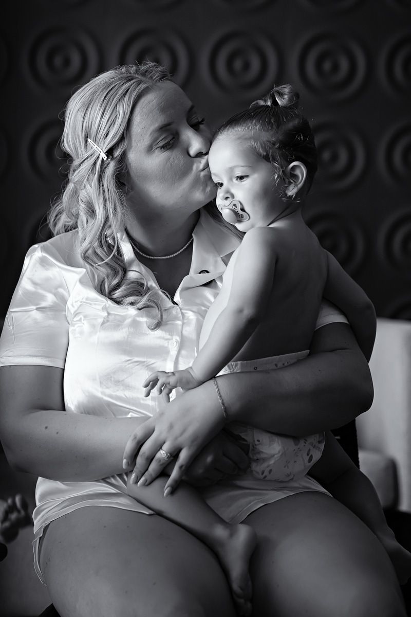 Black and white photo of a blonde woman kissing a toddler girl she holds in her lap indoors.