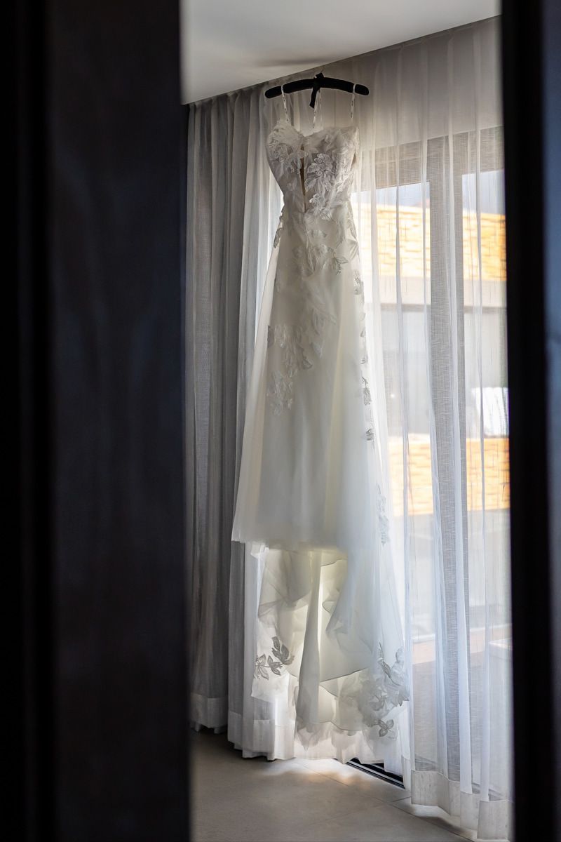 White lace wedding dress hanging on a hanger in front of sheer curtains by a bright window.