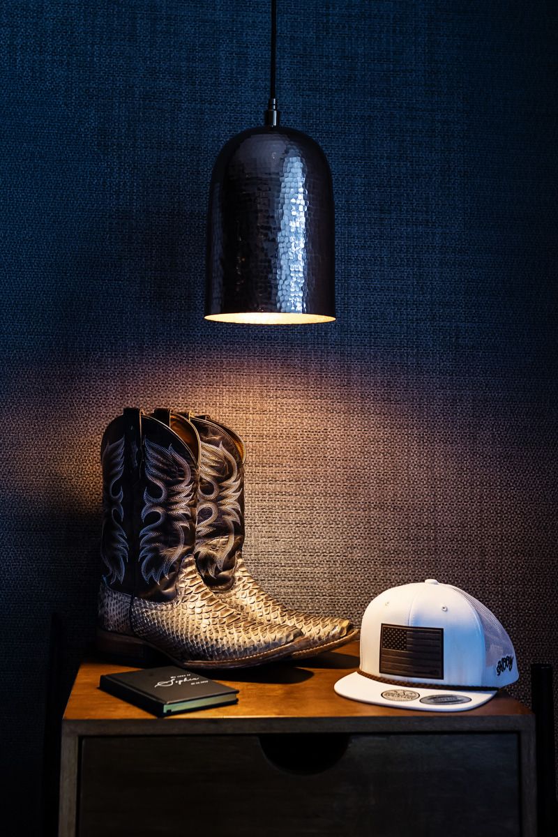 Cowboy boots, white snapback hat, and notebook on wooden shelf under pendant light against blue textured wall.