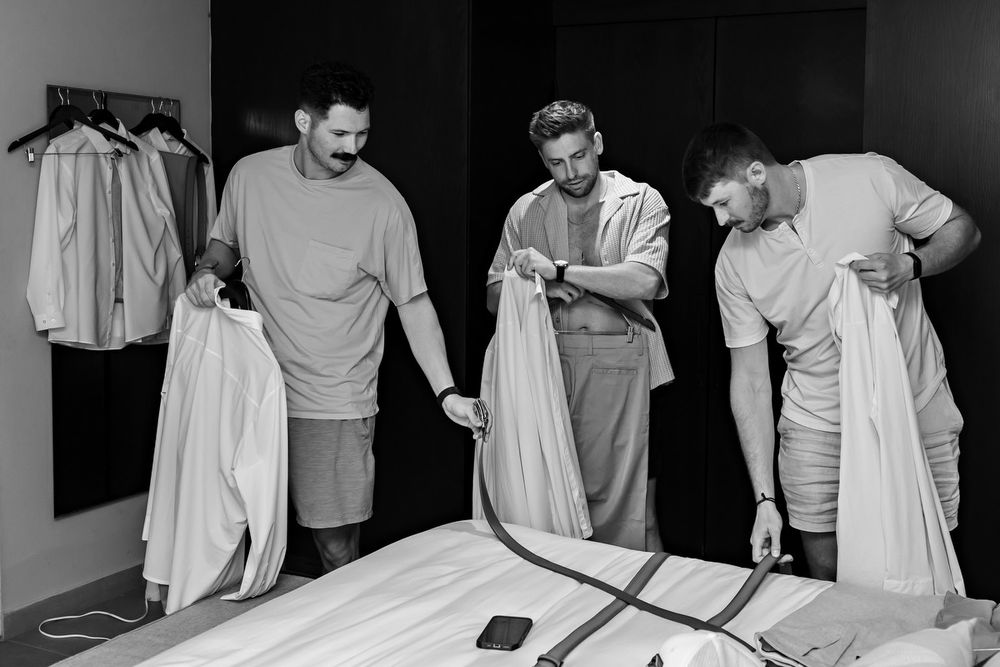 Three men examine white dress shirts in a fitting room with garment rack in black and white photo.