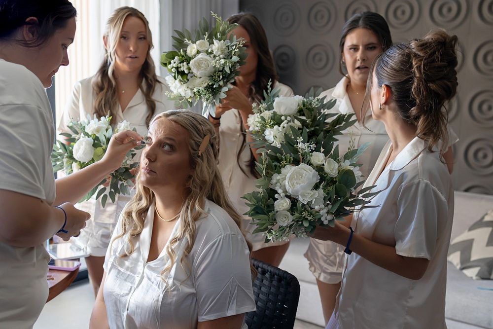 Bridesmaids in white robes holding bouquets of white flowers while helping bride prepare for wedding day.