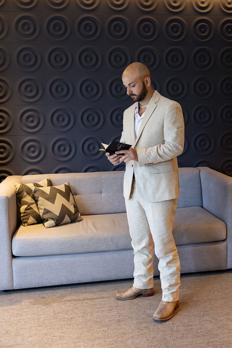 Bald bearded man in beige suit reading a book standing in modern hotel lobby with grey sofa.