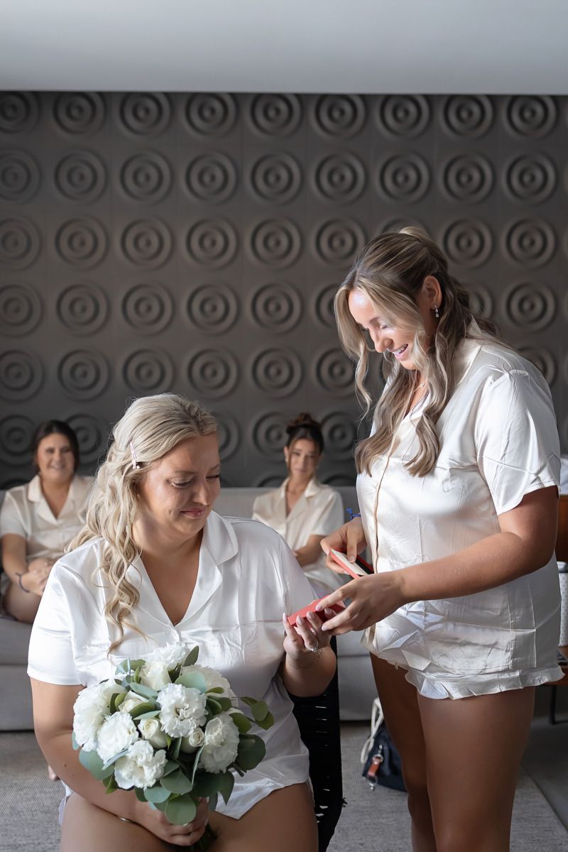 Bride holding white floral bouquet while bridesmaid shows her jewelry, all wearing white shirts during wedding preparations.