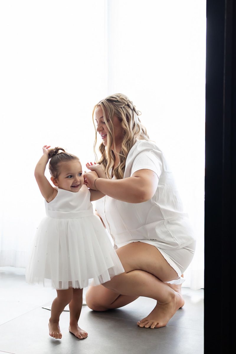 Mother and toddler daughter in matching white dresses share a tender moment together in a bright, airy room.