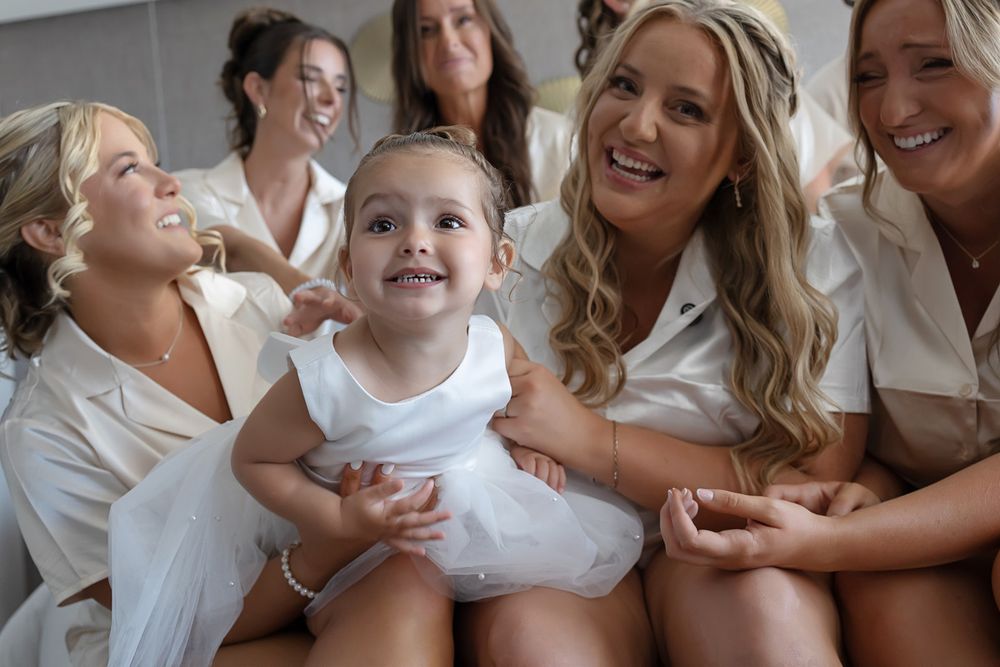 Bridesmaids in white robes smile and laugh with an adorable flower girl in a white dress during wedding preparations.