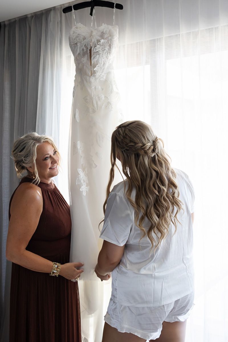 Two women admiring a white lace wedding dress hanging by a window in soft natural light.