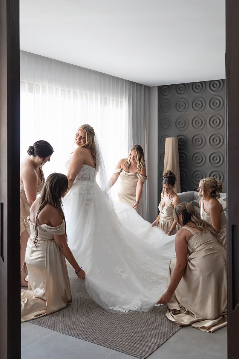 Bridesmaids in champagne gowns help bride in white lace wedding dress prepare in elegant hotel room.
