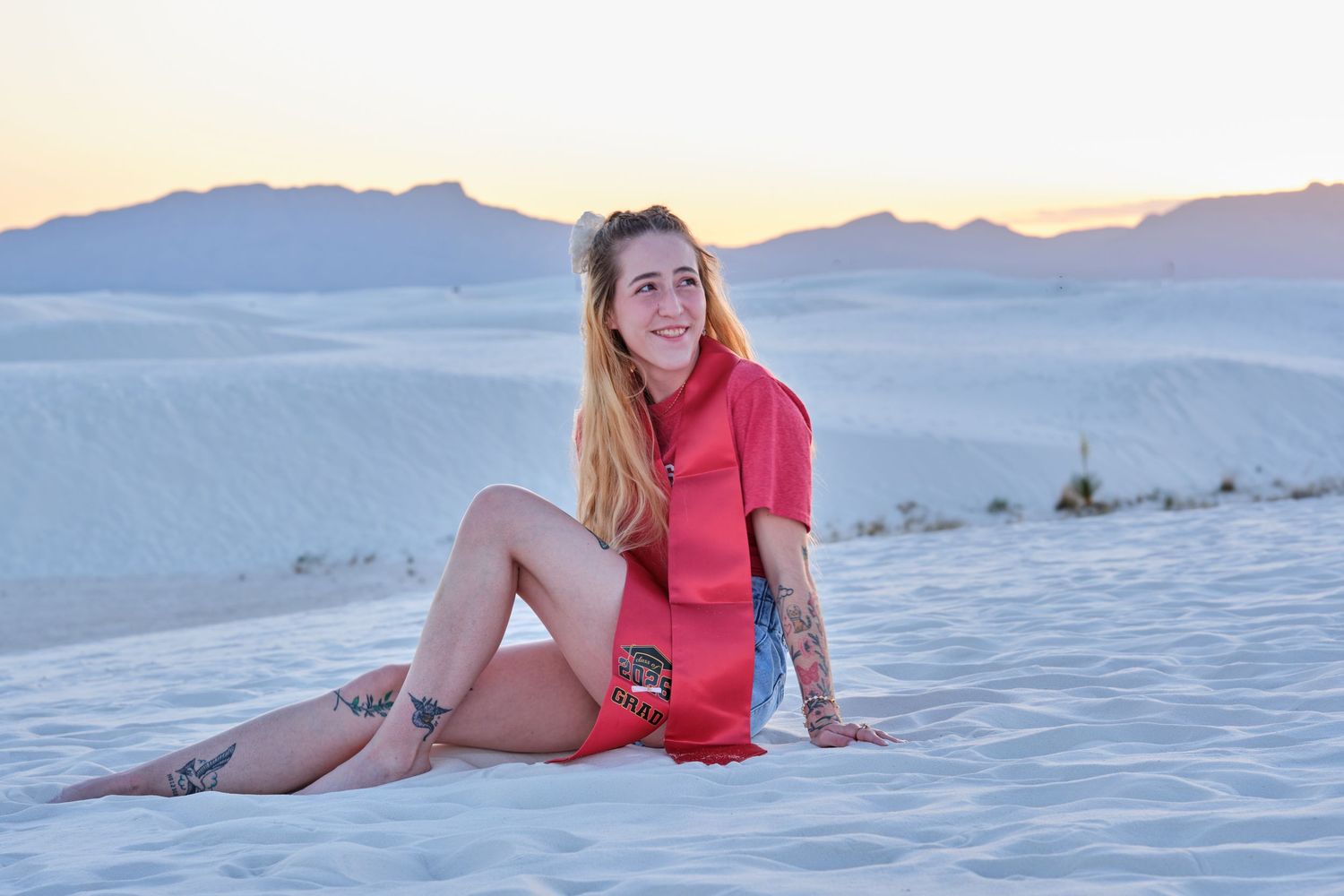 Woman in red outfit sitting on white sand dunes at sunset with mountains in the background.