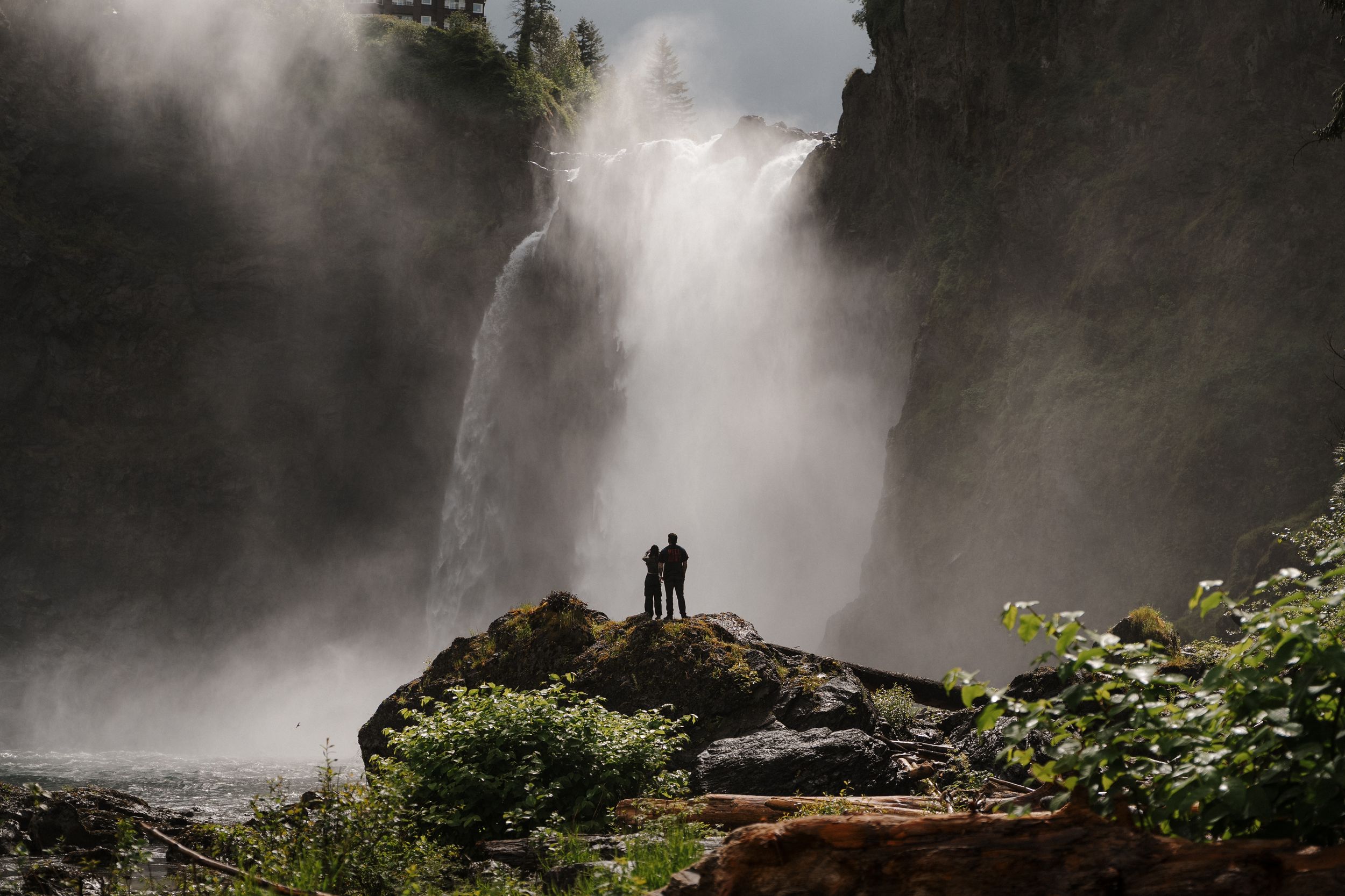 Two people stand on a rock before a massive misty waterfall surrounded by lush green vegetation in a dramatic landscape.