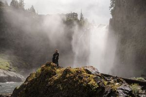Person standing on mossy rocks before a massive misty waterfall in a lush green forest landscape.
