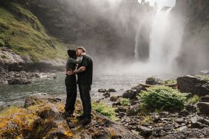 Couple embracing romantically in front of a misty waterfall surrounded by lush greenery and rocky terrain.