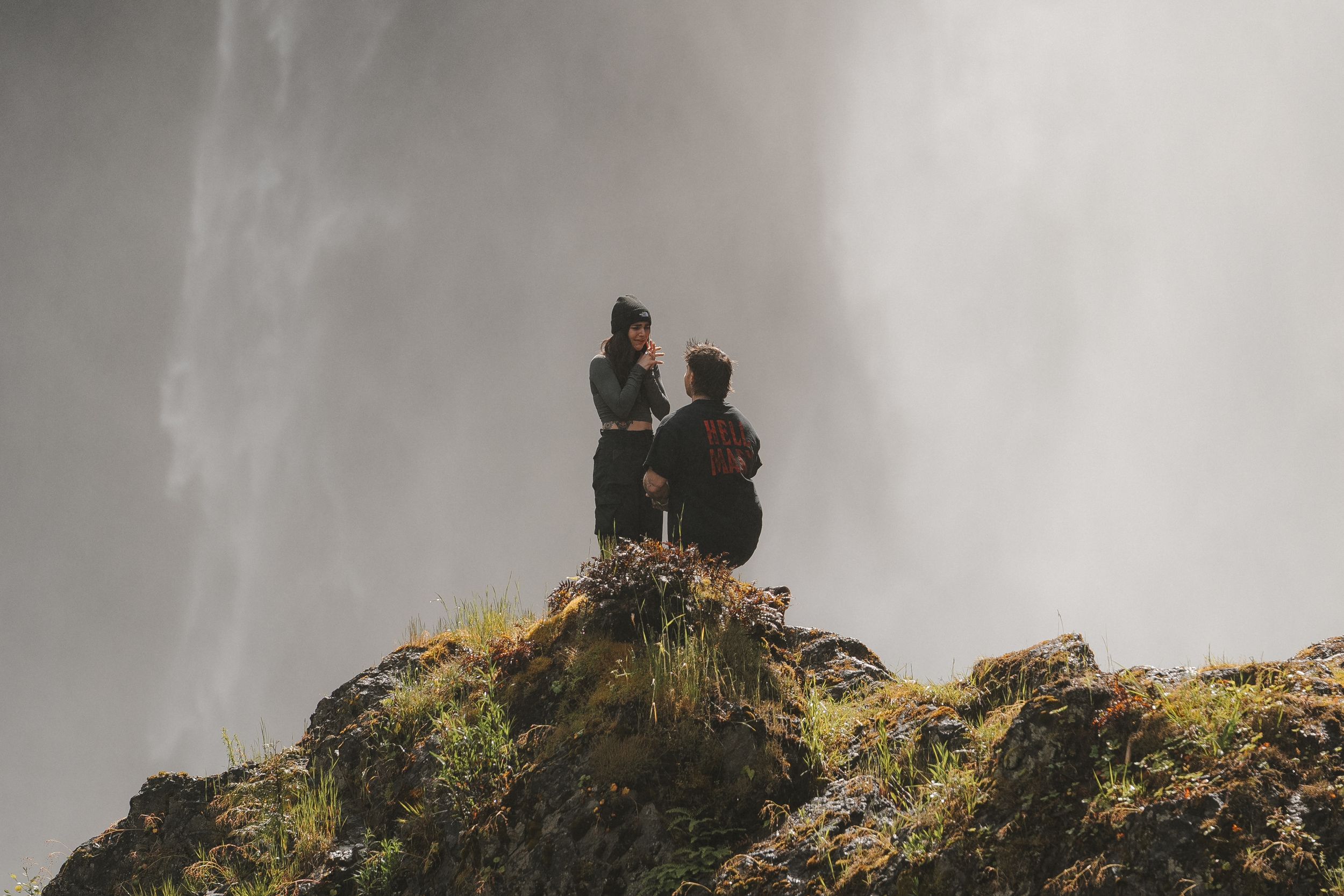 Two people stand on a rocky cliff overlooking a misty waterfall in a dramatic outdoor landscape.