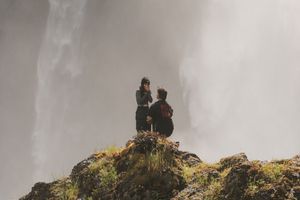 A person proposing on a rocky cliff with a dramatic misty waterfall backdrop in a scenic outdoor setting.