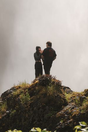 Two hikers stand on a misty rocky hilltop surrounded by lush greenery, facing each other in conversation.