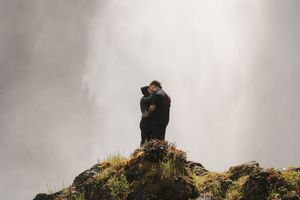 Couple embracing romantically on a misty cliff edge surrounded by lush greenery and dramatic foggy waterfall backdrop.