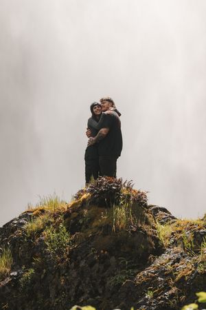 Couple embracing on a misty rocky hilltop surrounded by wild grass and fog in a romantic outdoor setting.