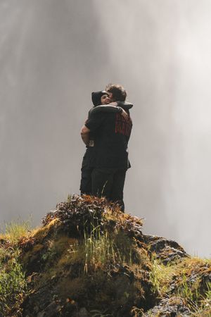 Couple embracing on a misty rocky outcrop near a waterfall in a dramatic outdoor landscape.
