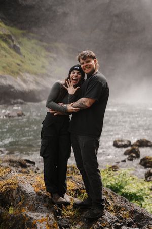 Happy couple posing by a misty waterfall, woman showing engagement ring while man hugs her.