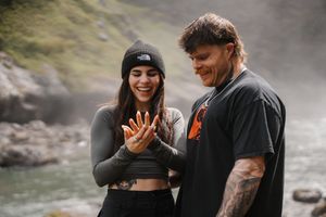 A tattooed couple smiling outdoors near a misty waterfall, woman wearing North Face beanie examining her hands.
