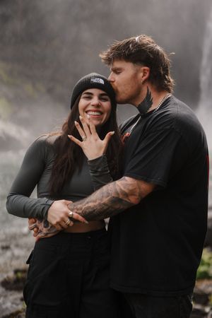 Couple celebrating engagement near waterfall, woman showing ring while man kisses her cheek, both wearing black.