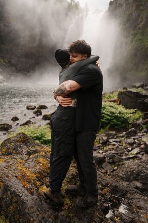Couple embracing tightly near misty waterfall on rocky terrain, both dressed in black outdoor attire.