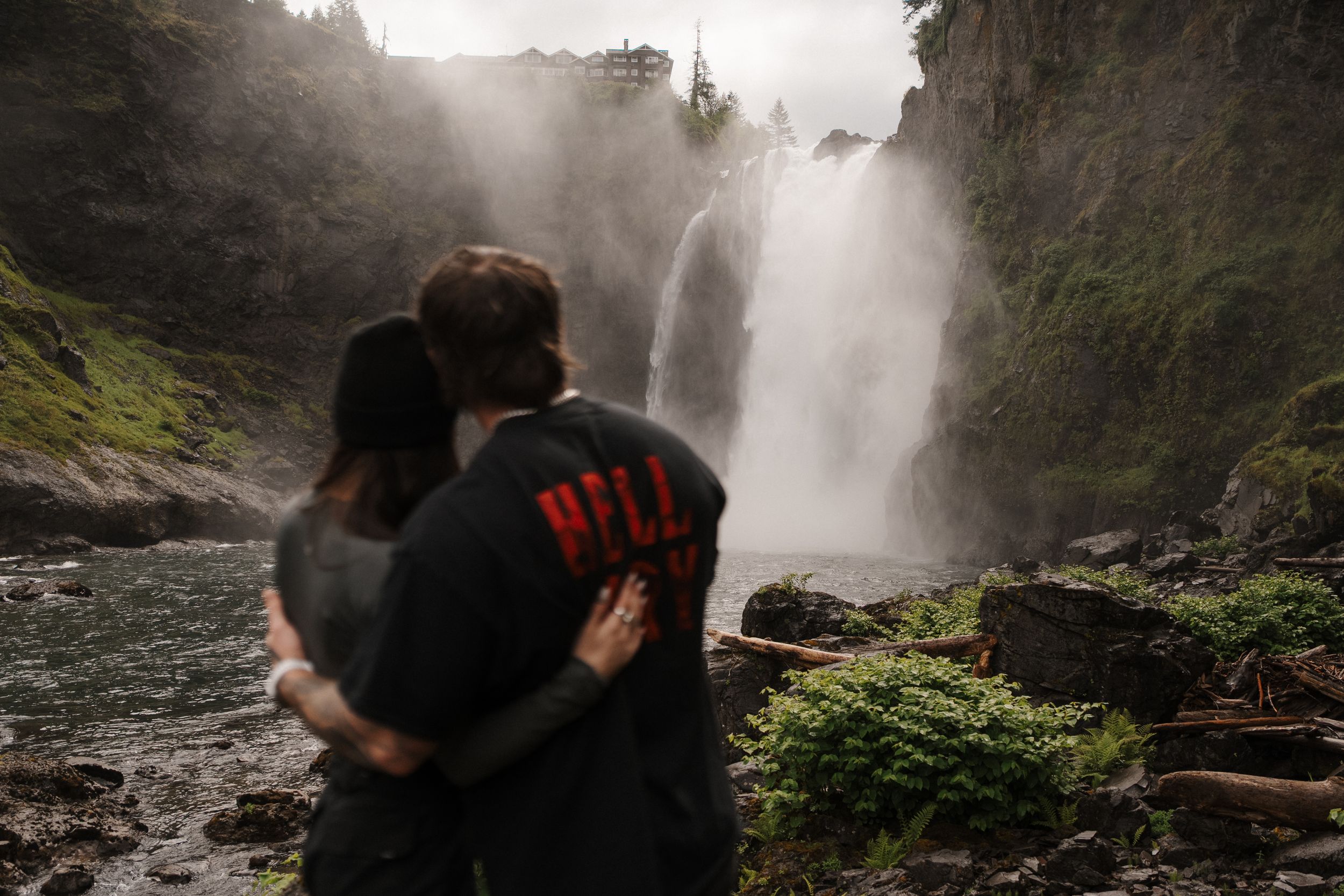 Couple embracing before a misty waterfall surrounded by lush greenery, romantic outdoor photography.