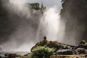 Person standing on a rock before a massive waterfall with mist rising, a building visible on the cliff above.