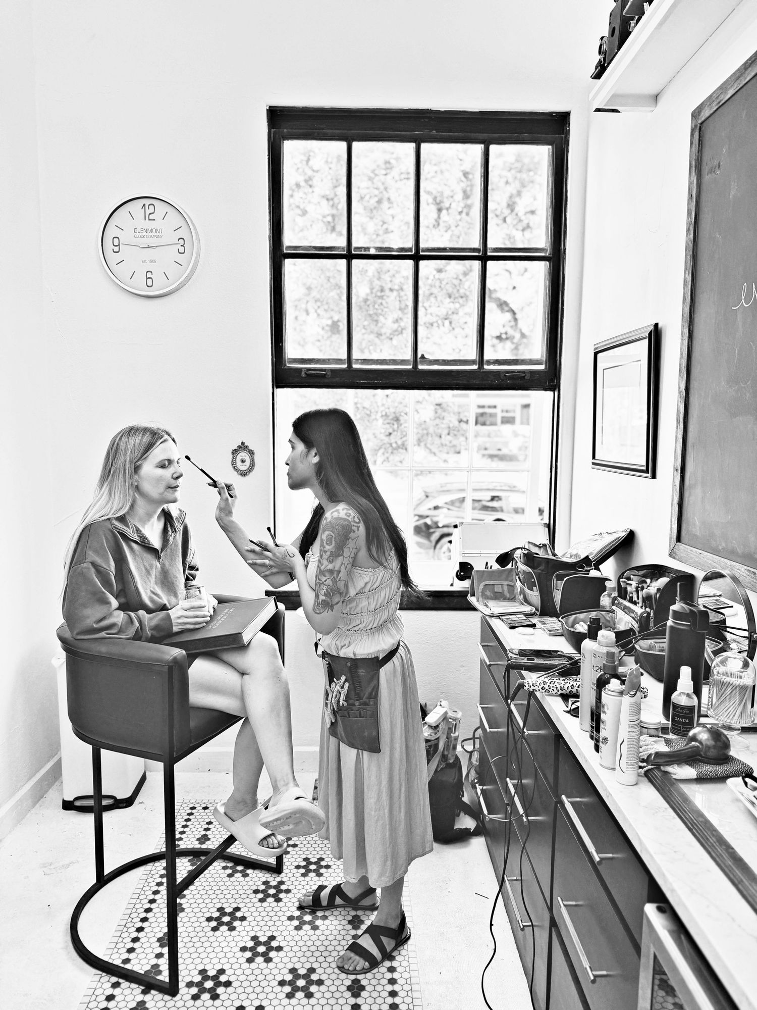 A makeup artist applies cosmetics to a seated blonde woman near a window in a bright studio with a patterned tile floor.