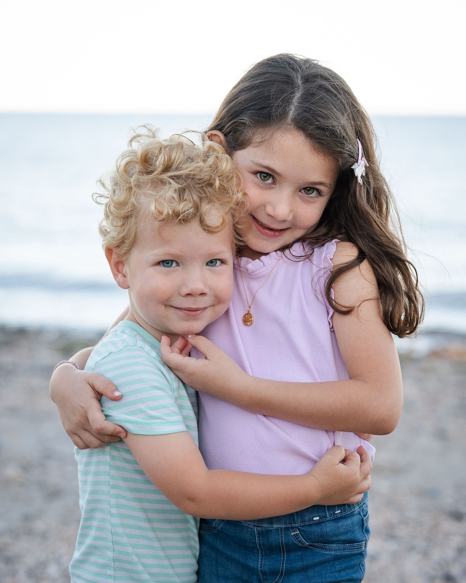Two young children holding hands and running on a sandy beach near the ocean on a cloudy day.
