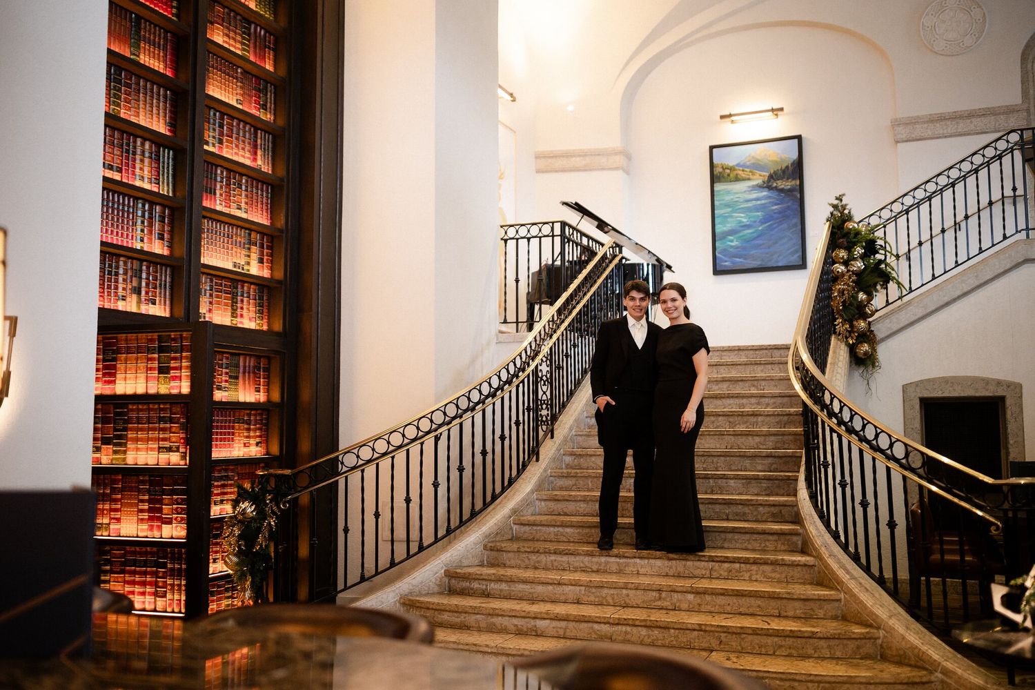 Groom and a loved one in black on the grand staircase beside a wall of books at a Fairmont Banff Springs winter wedding