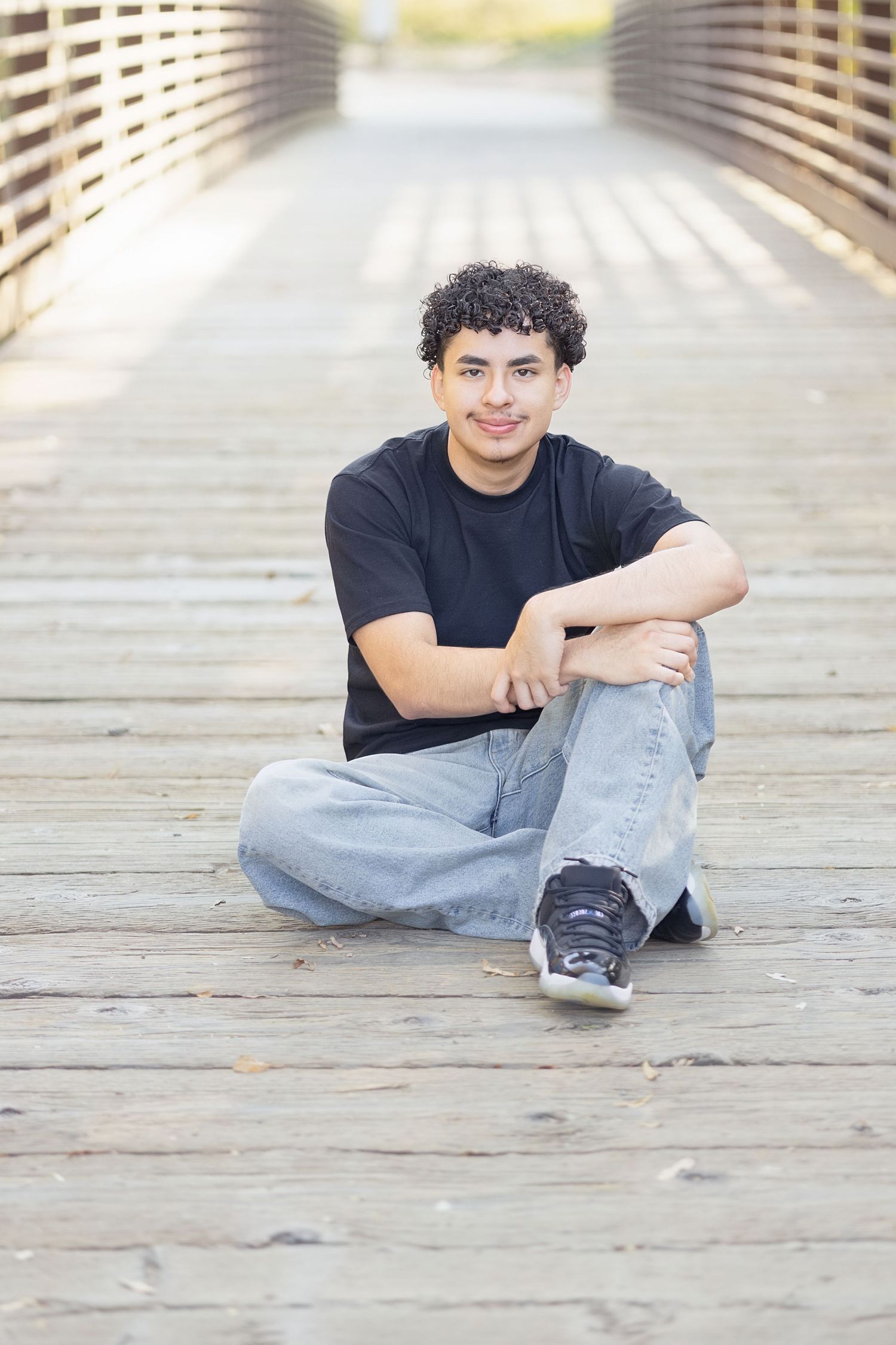 Young man with curly hair sitting cross-legged on a wooden bridge, wearing a black t-shirt and jeans, smiling.