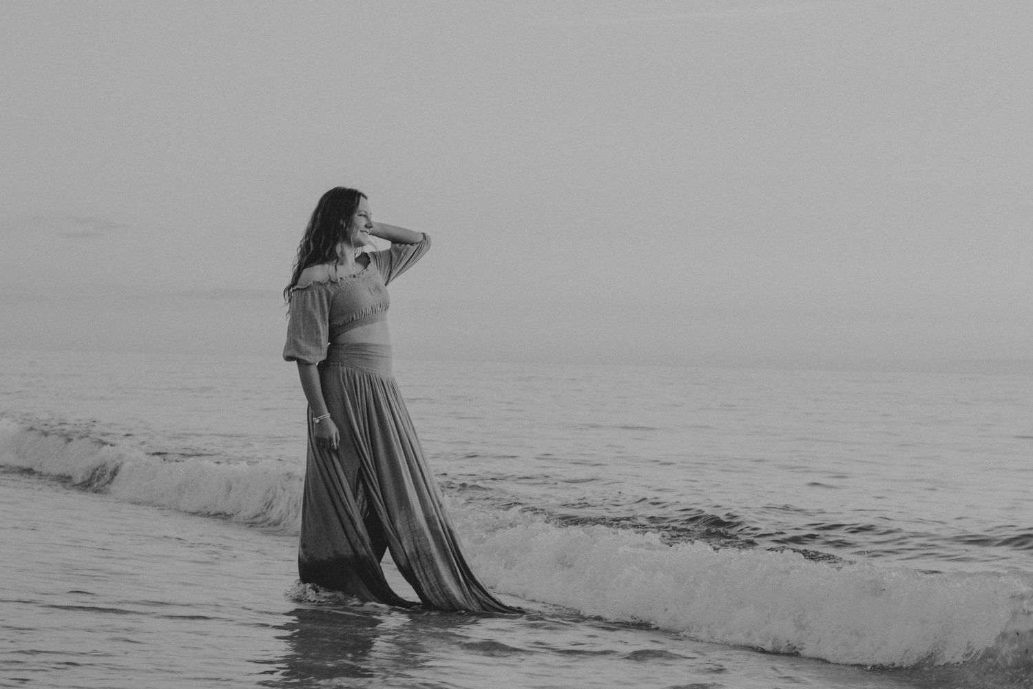 A woman in a flowing dress stands gracefully at the ocean shoreline in a serene black and white beach photo.