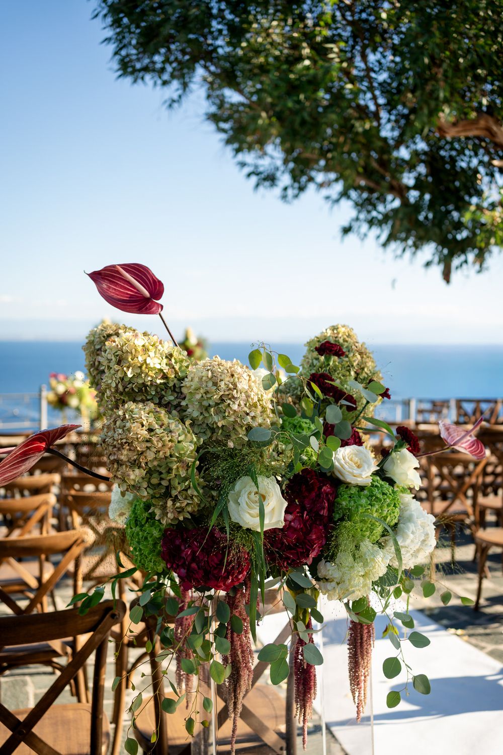 Elegant outdoor wedding ceremony floral arrangement with burgundy anthuriums, white roses, and hydrangeas overlooking the ocean.