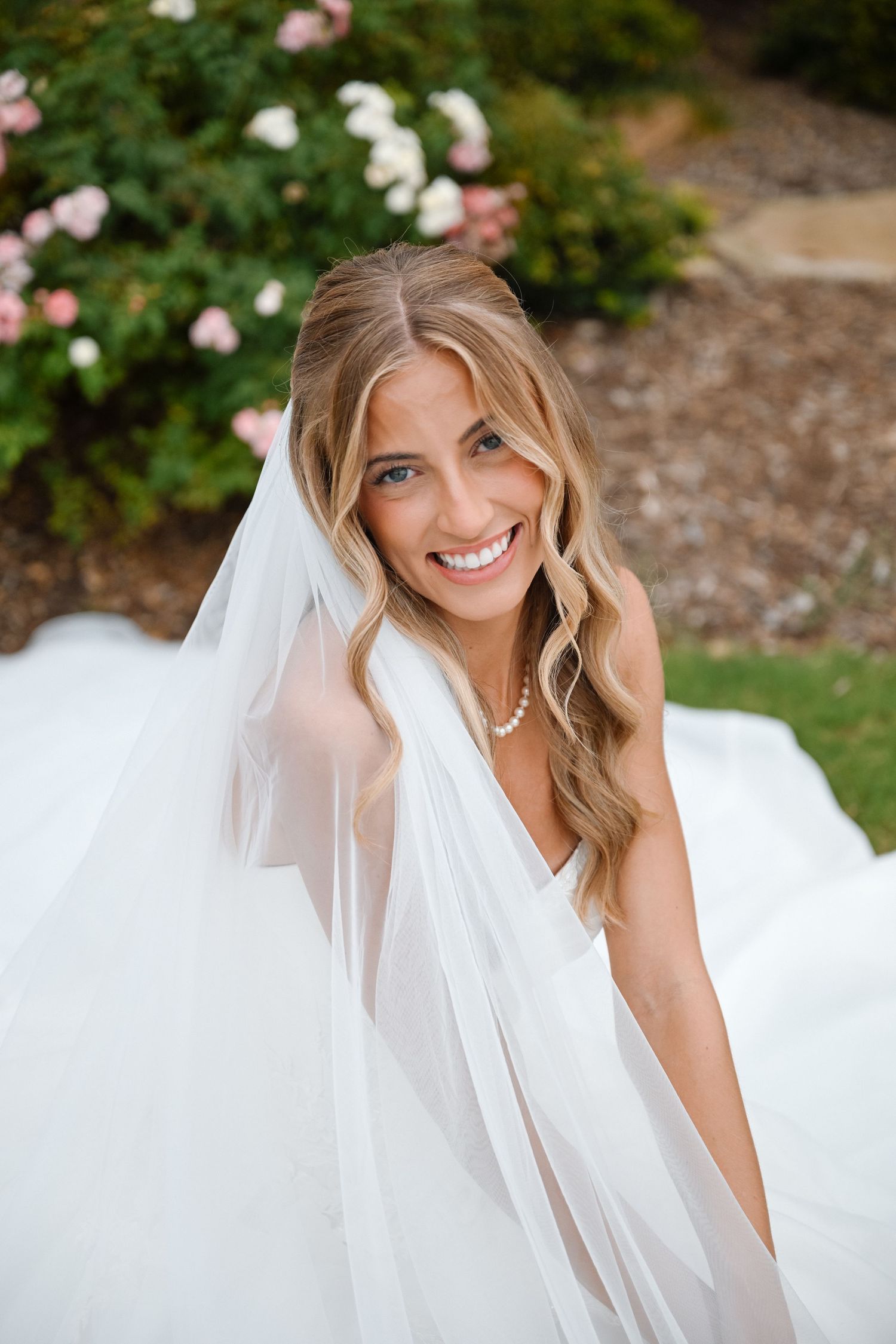 Smiling bride in white gown and veil wearing pearl necklace, posing outdoors with blooming flowers in background.