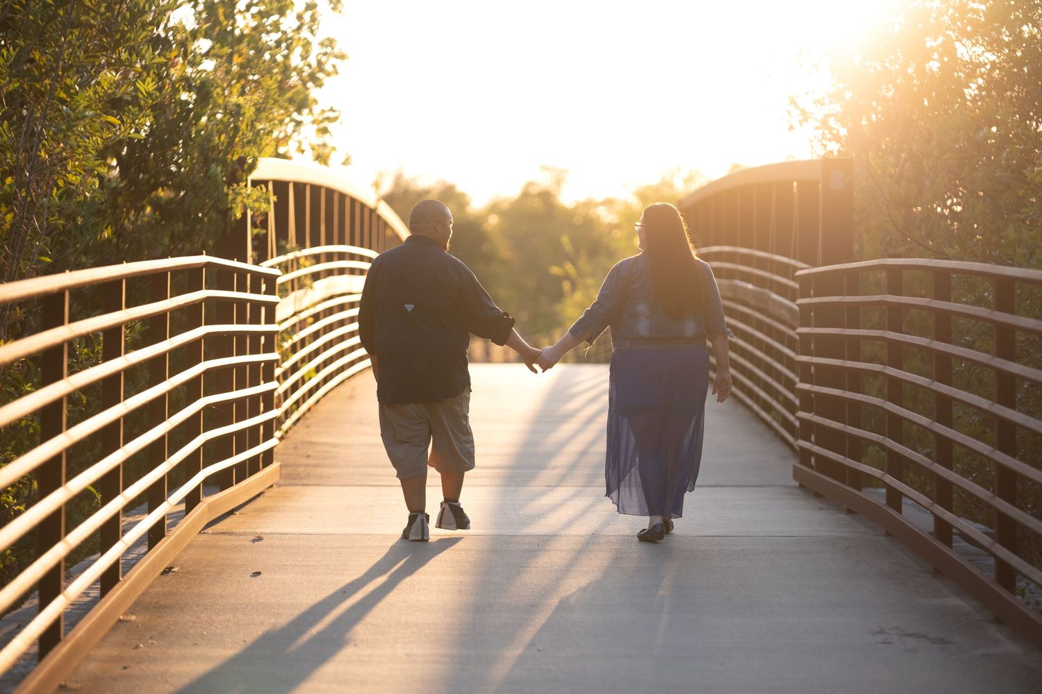 Couple holding hands walking on a sunlit bridge at golden hour, surrounded by lush green trees.