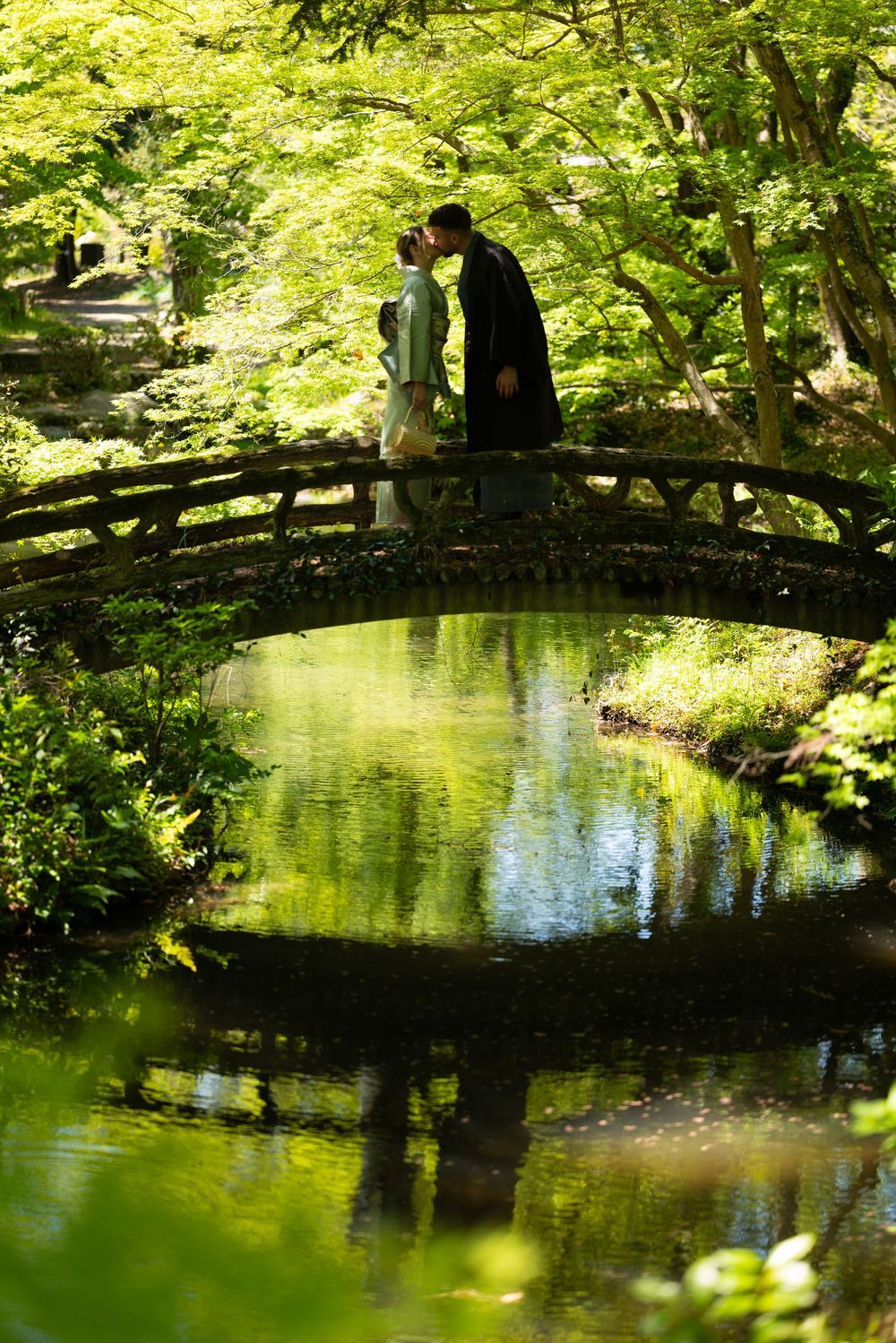 Couple in traditional Japanese attire shares a quiet moment on an arched wooden bridge over a calm pond surrounded by lush green trees.